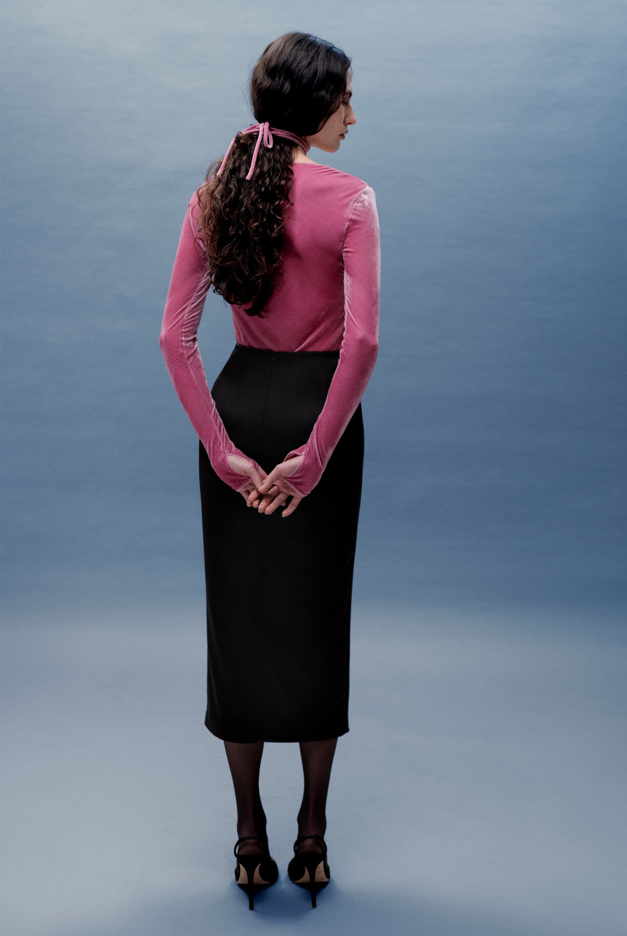 Woman wearing a pink long-sleeve Massima top and black skirt against a gray background