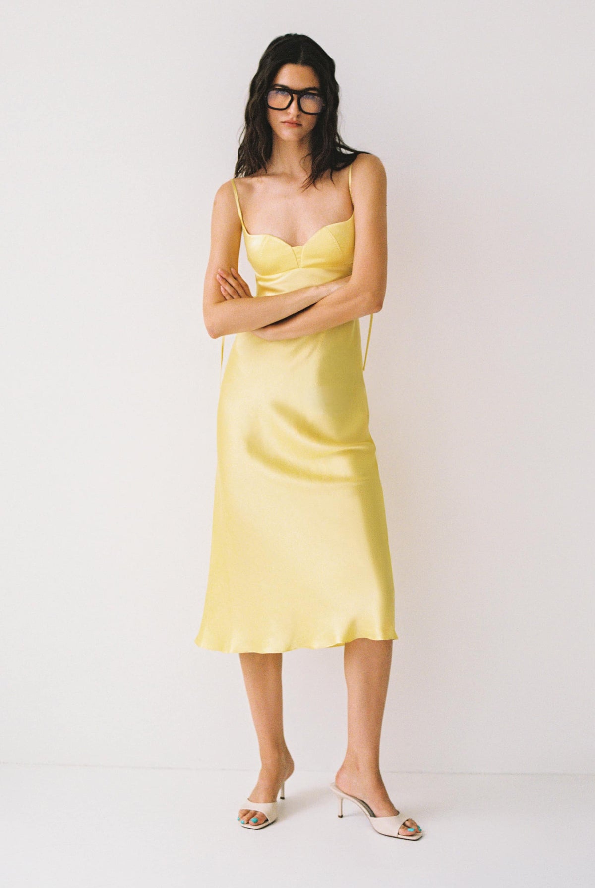 A woman modeling a pale yellow satin slip Isolde dress with thin straps, paired with white heeled mule sandals and dark sunglasses, against a plain white studio background
