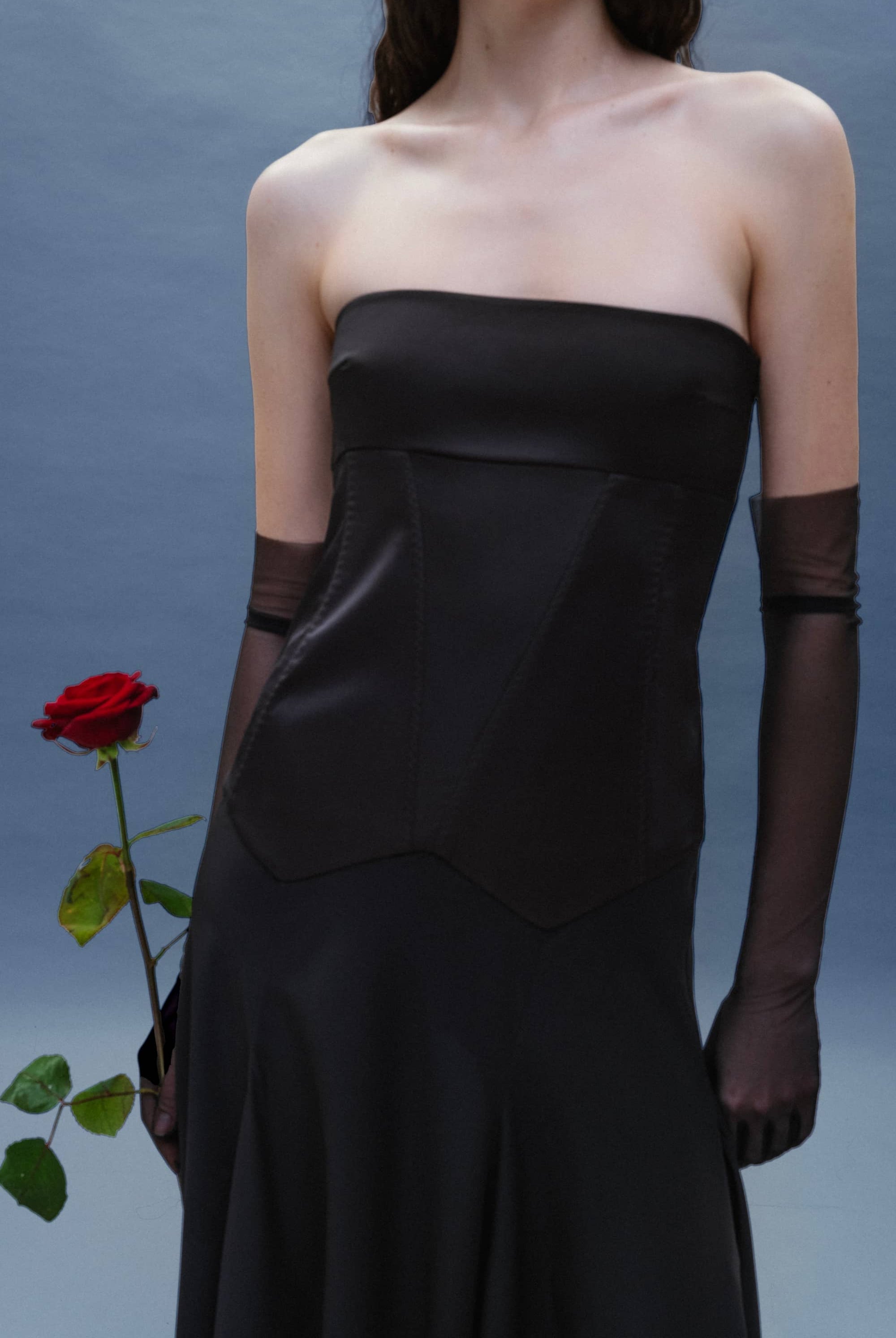 Woman in a black strapless Ariella dress holding a red rose against a gray background