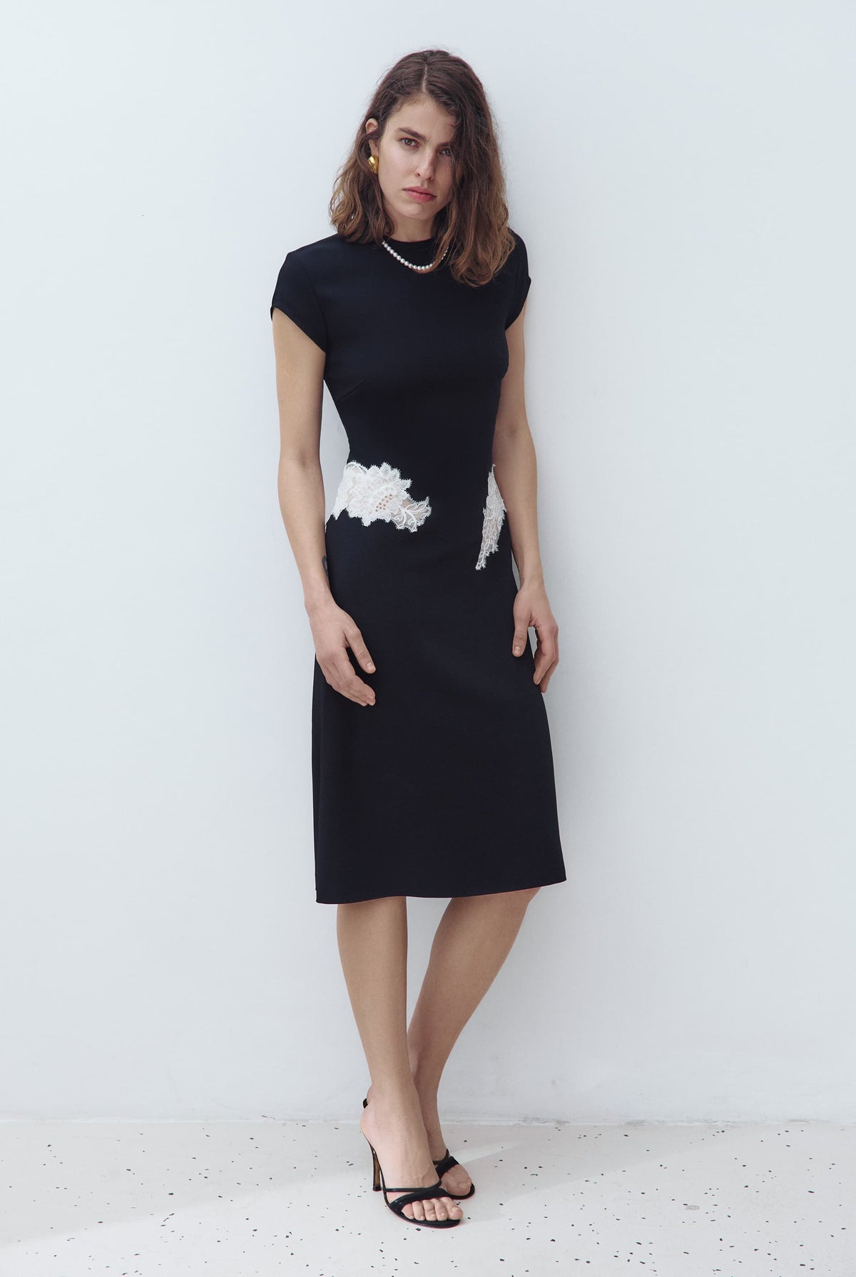 A woman modeling a black knee-length Eleni dress with short cap sleeves and a white textured floral applique detail at the waist, standing against a plain light gray wall