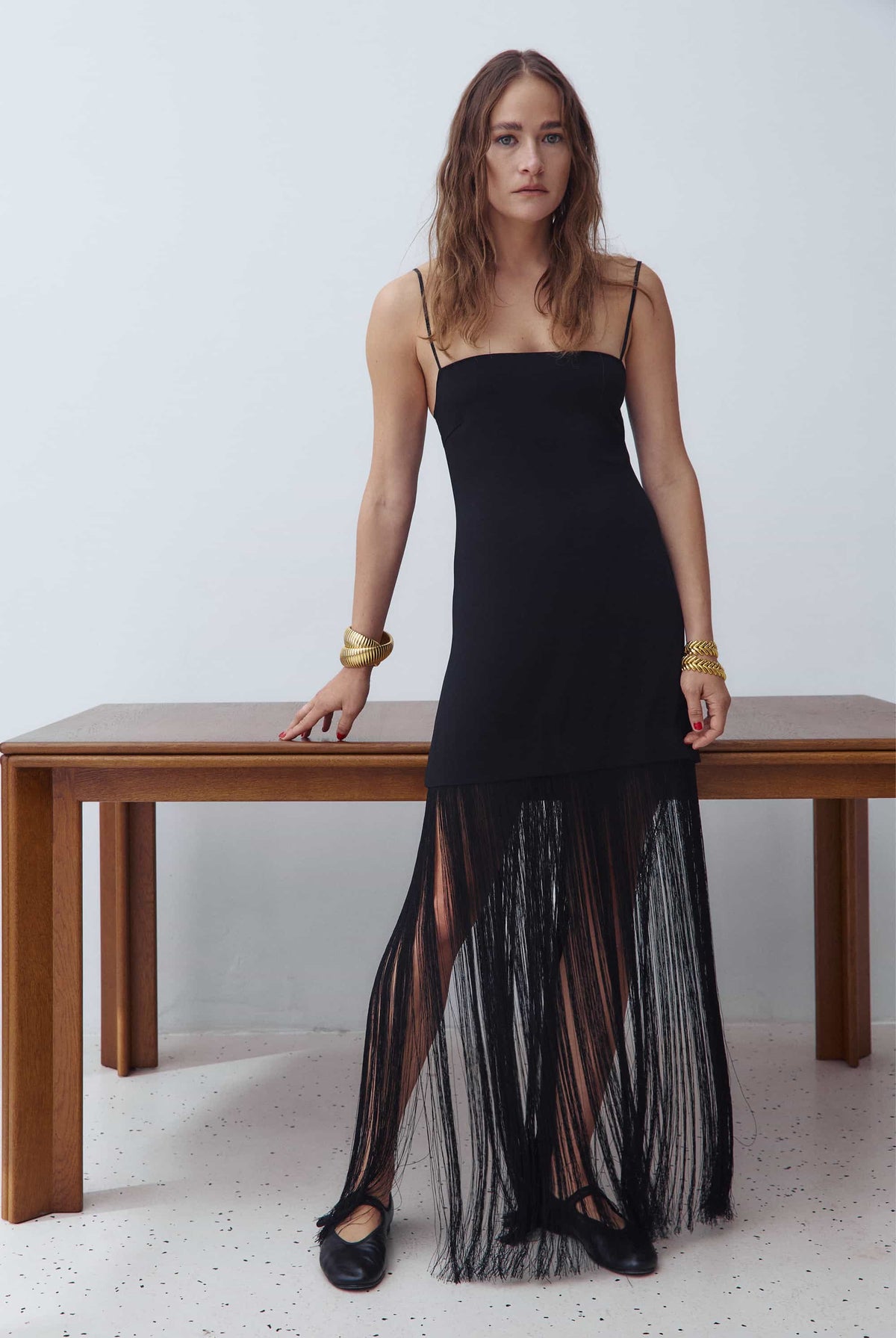 A woman models a black spaghetti-strap maxi Evangelia dress with a solid upper bodice and a sheer, fringed tulle hem, standing next to a wooden table against a white wall