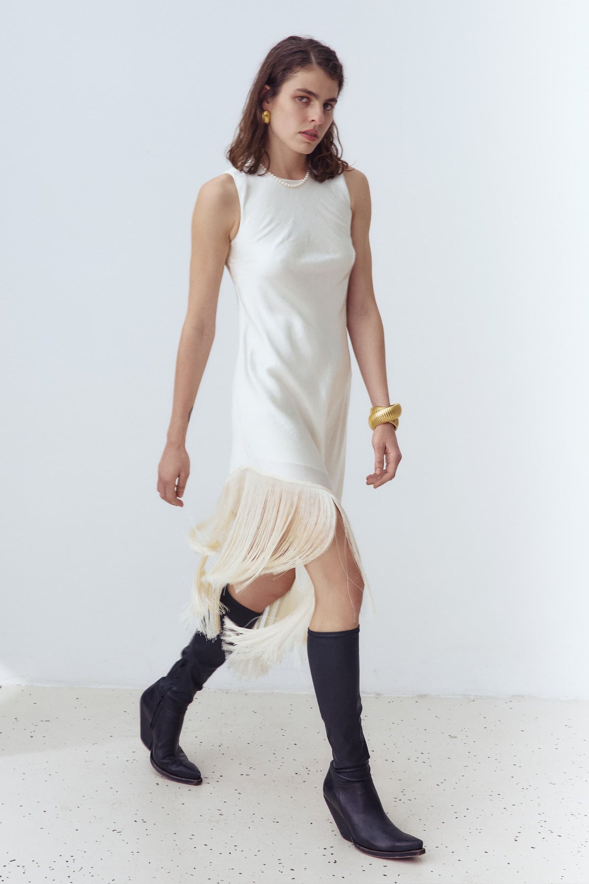 A woman modeling a sleeveless ivory Amiyra dress with a pleated fringe hem, paired with black knee-high boots, against a clean white studio background