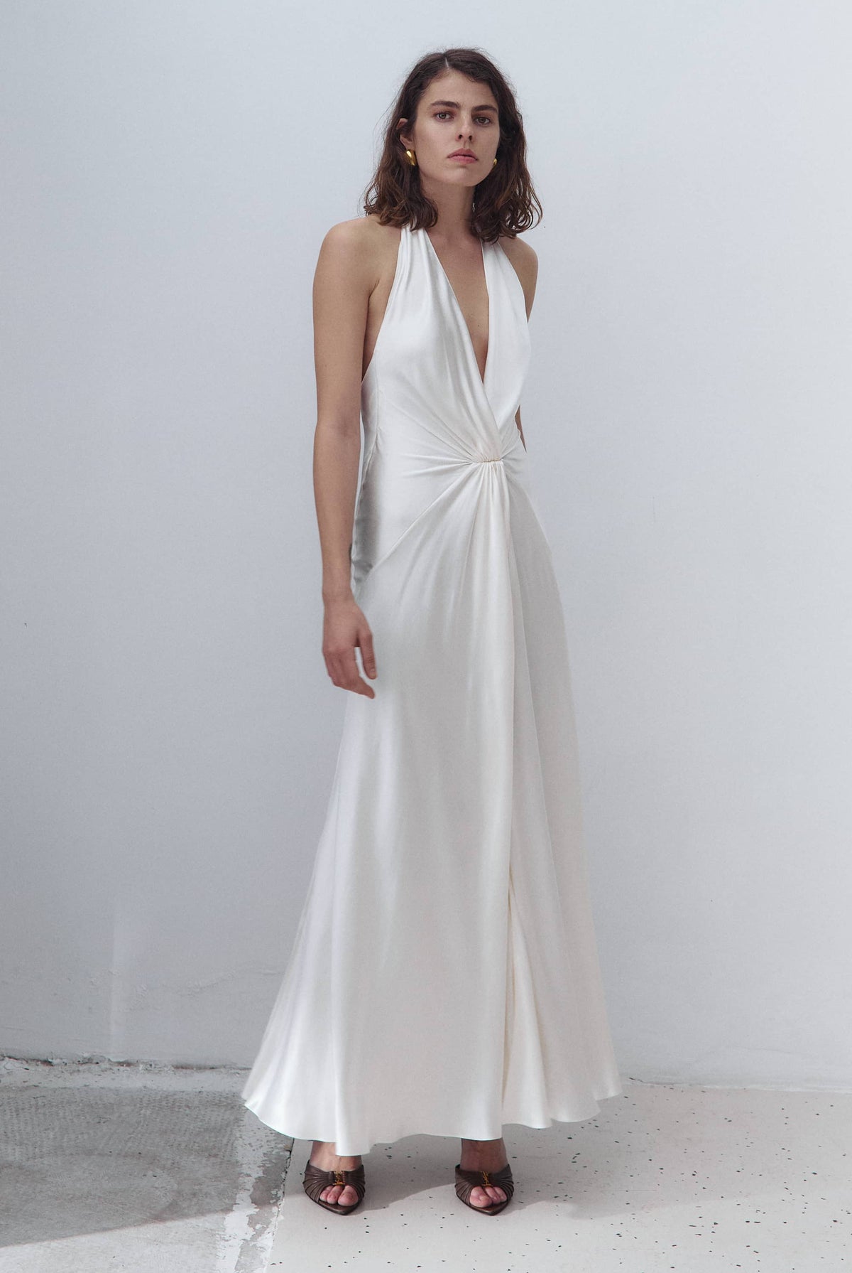 A woman modeling a floor-length white halter Ruth dress with a deep V-neckline and a gathered twist detail at the waist, standing against a plain light gray wall