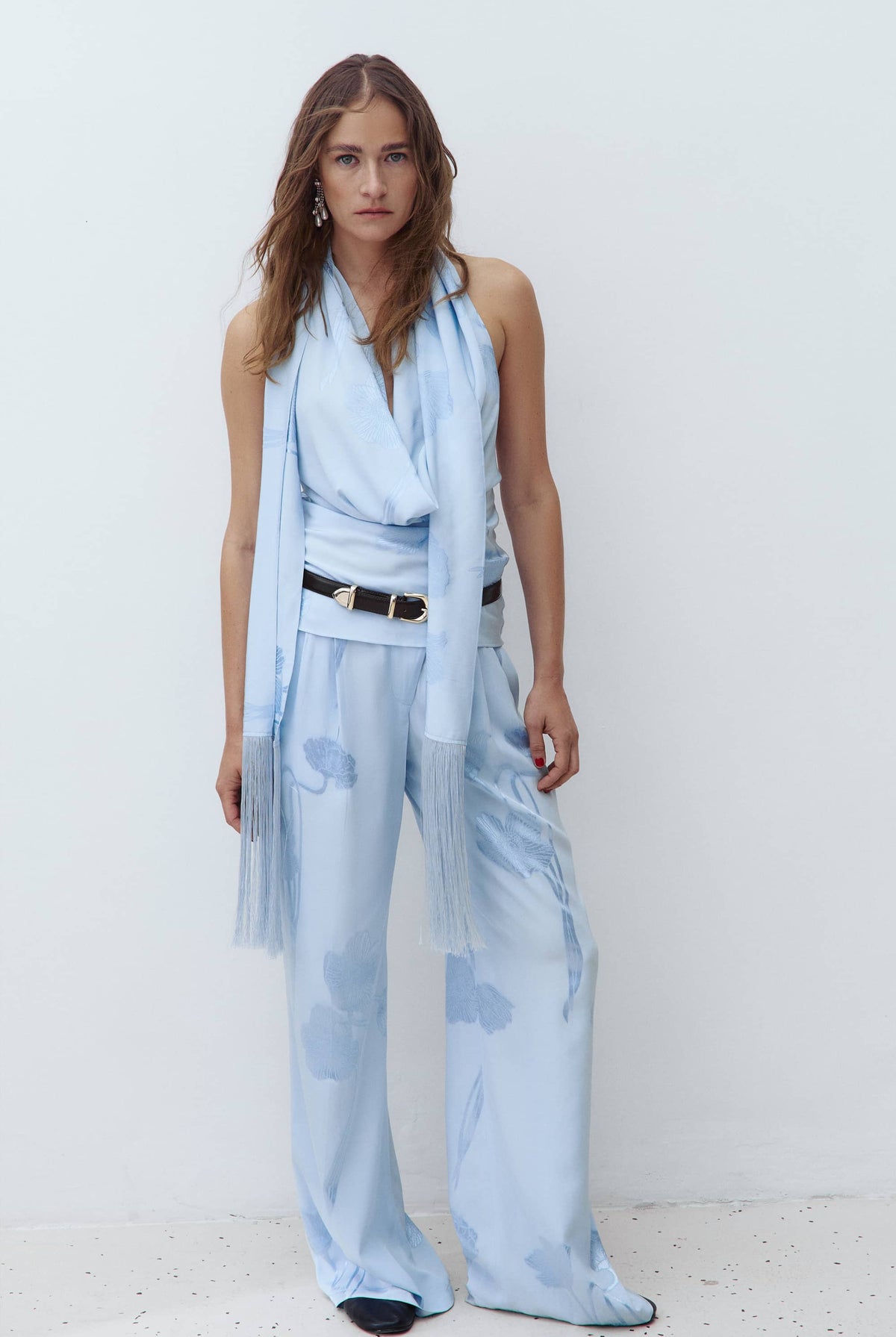 A woman modeling a light blue sleeveless wrap-style Scarlet top and matching wide-leg pants with a sheer tulle scarf, styled with a black belt, against a plain white studio background