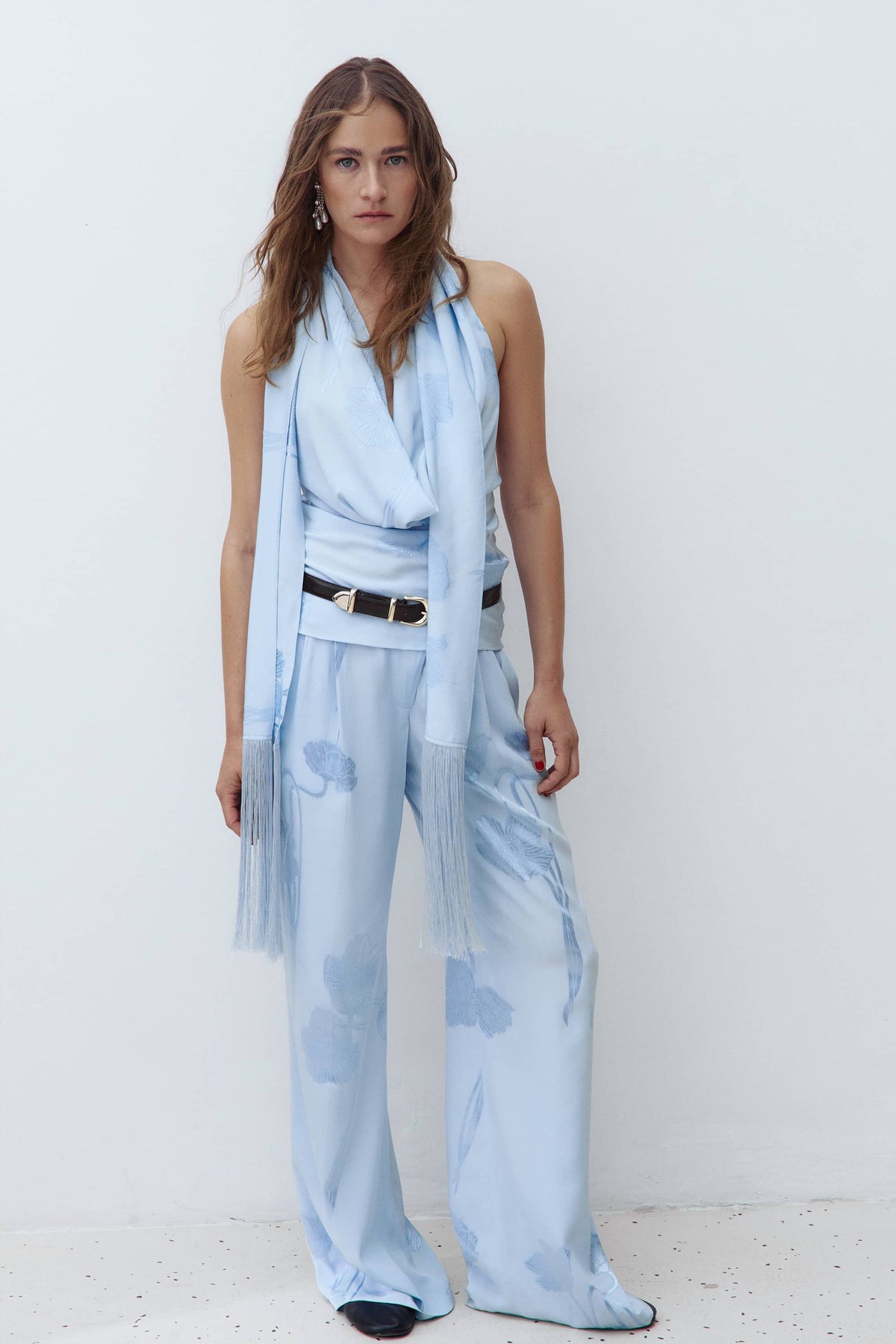 A woman modeling a light blue sleeveless wrap-style Scarlet top and matching wide-leg pants with a sheer tulle scarf, styled with a black belt, against a plain white studio background