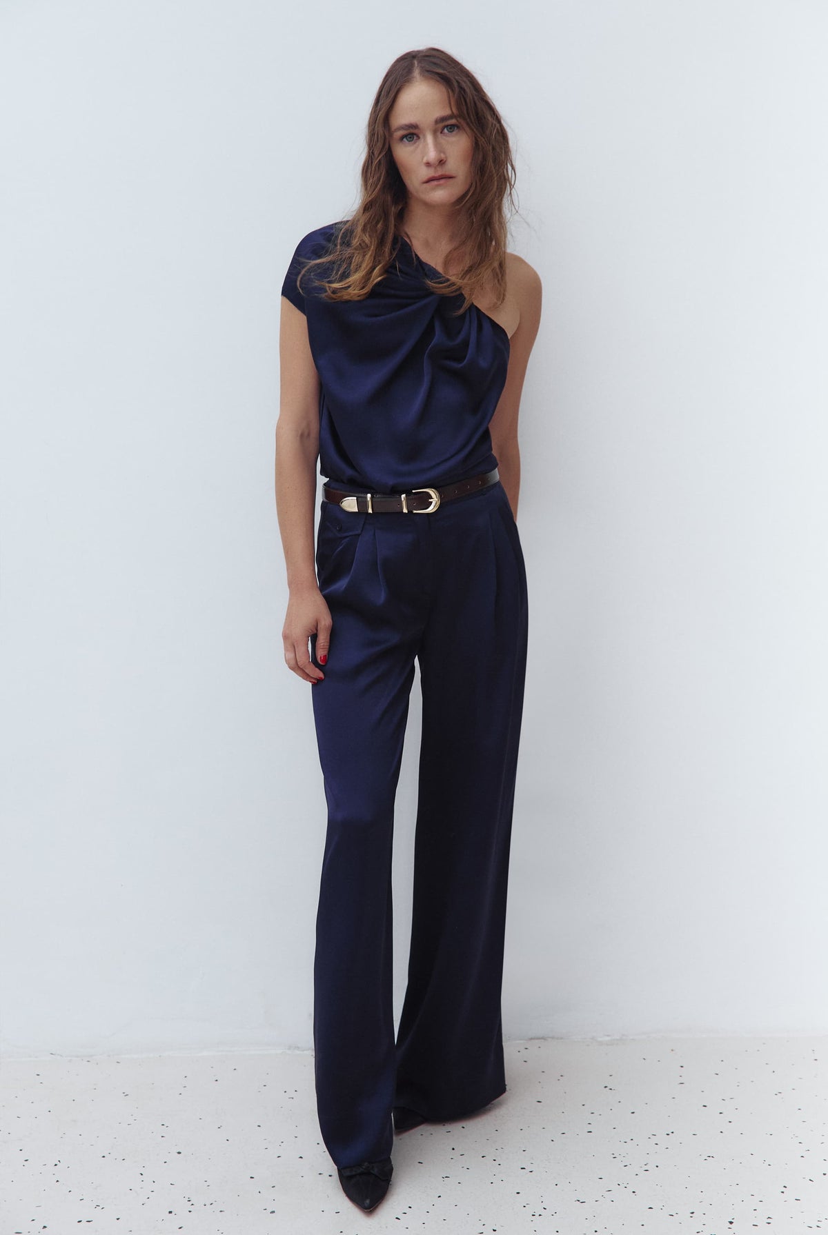 A woman modeling a dark navy blue one-shoulder draped Paris top and matching navy wide-leg Katerina trousers, styled with a brown belt and gold bracelets against a light gray studio background