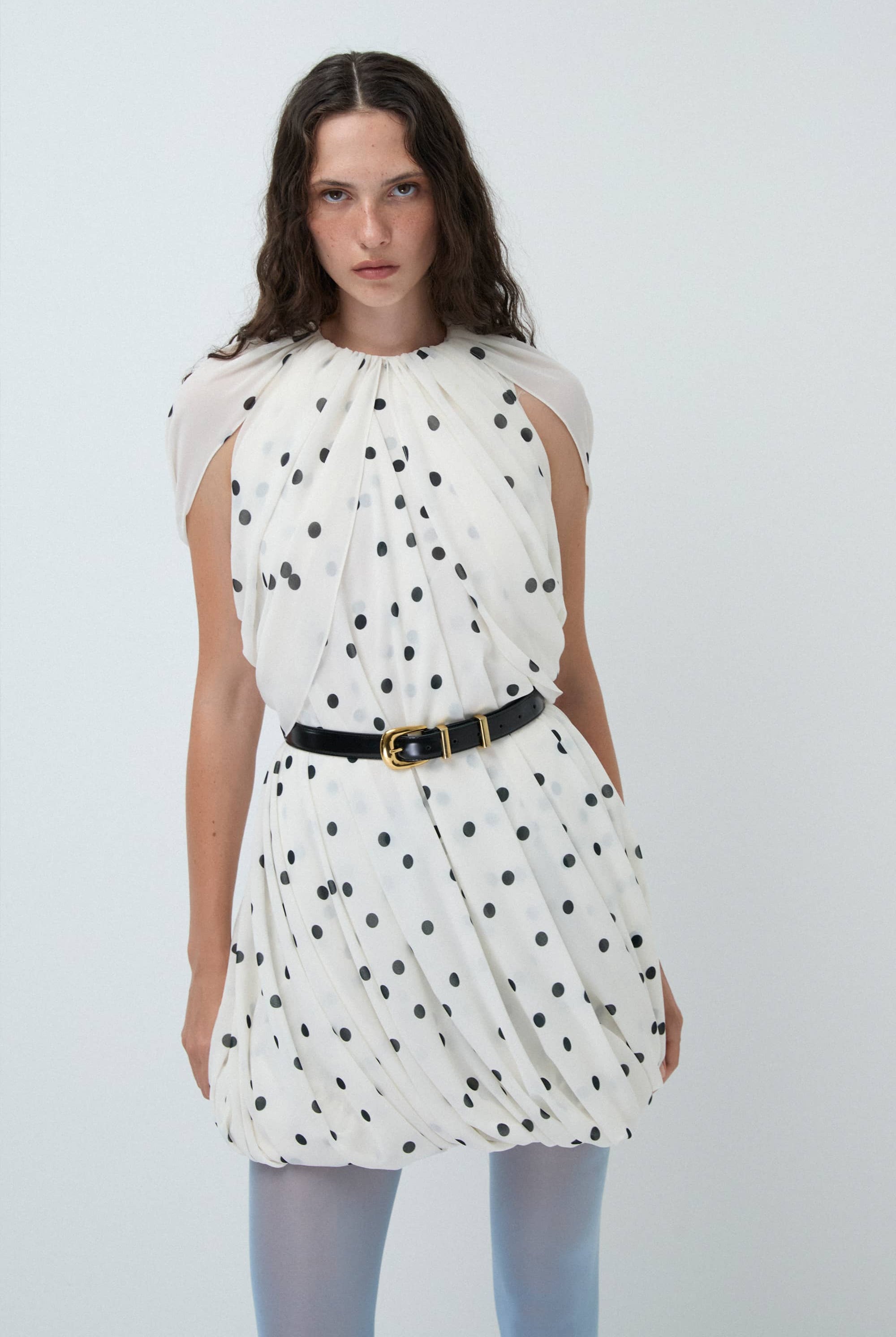 Woman wearing a white Mio dress with black polka dots and a black belt on a light gray background, front view