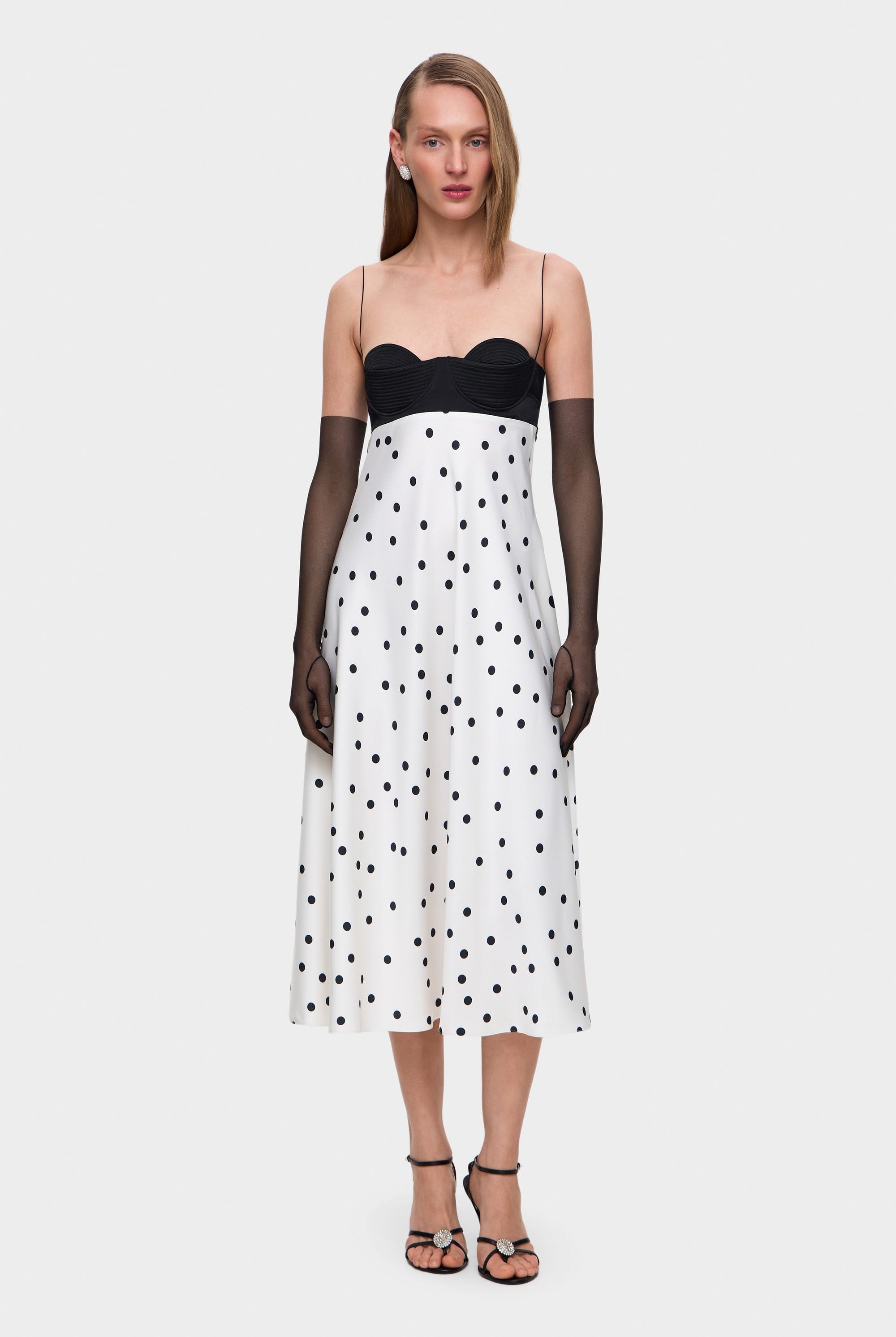 Woman wearing a black and white polka dot Erika dress with thin straps and black gloves on a white background, front view