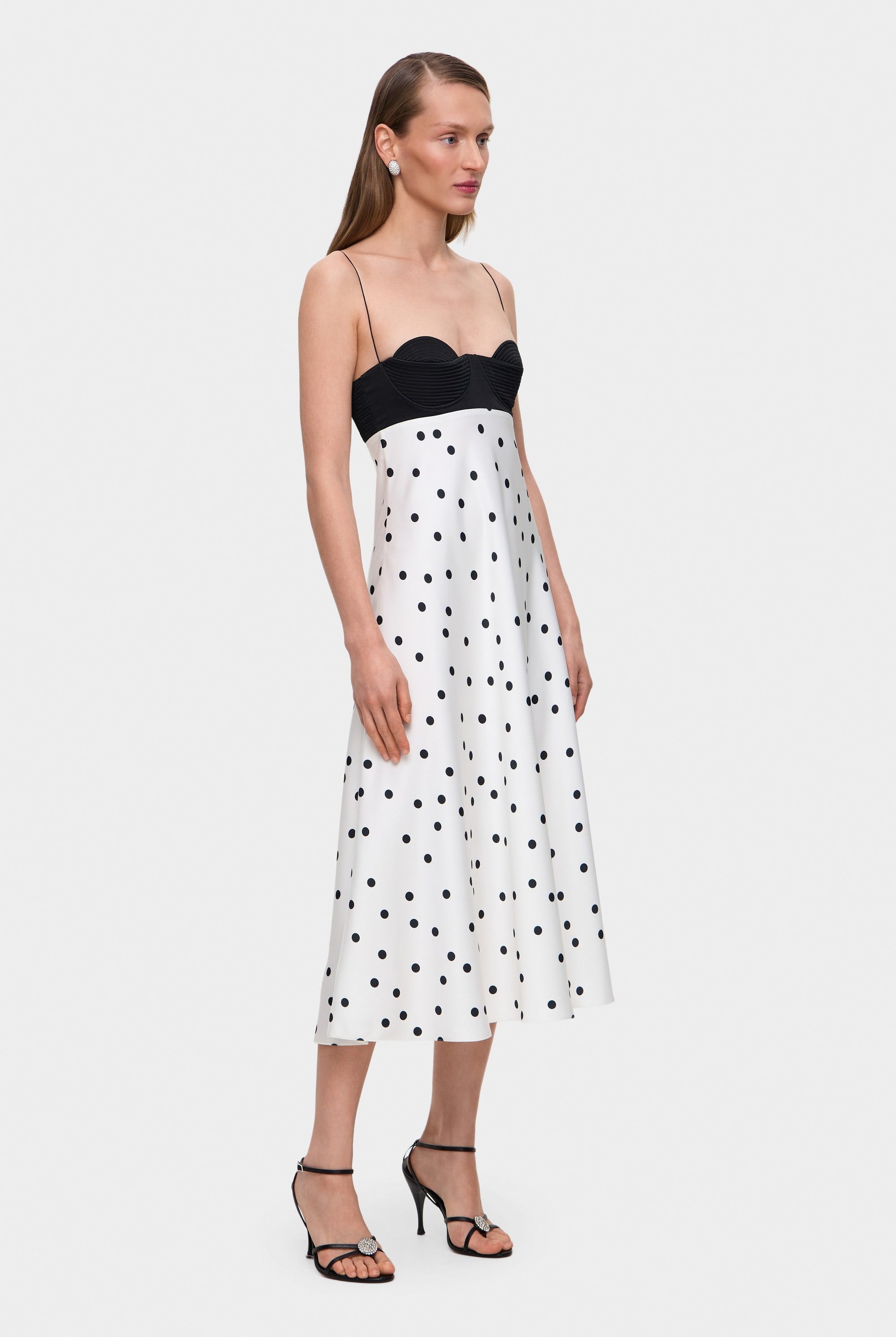 Woman wearing a black and white polka dot Erika dress with thin straps on a white background, side view