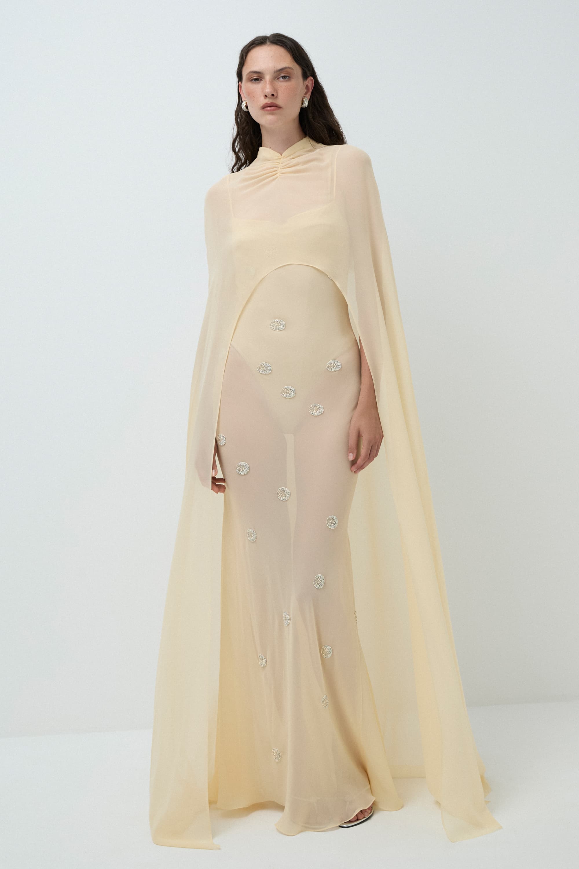 Woman wearing a buttermilk Hina dress with a cape on a white background, front view