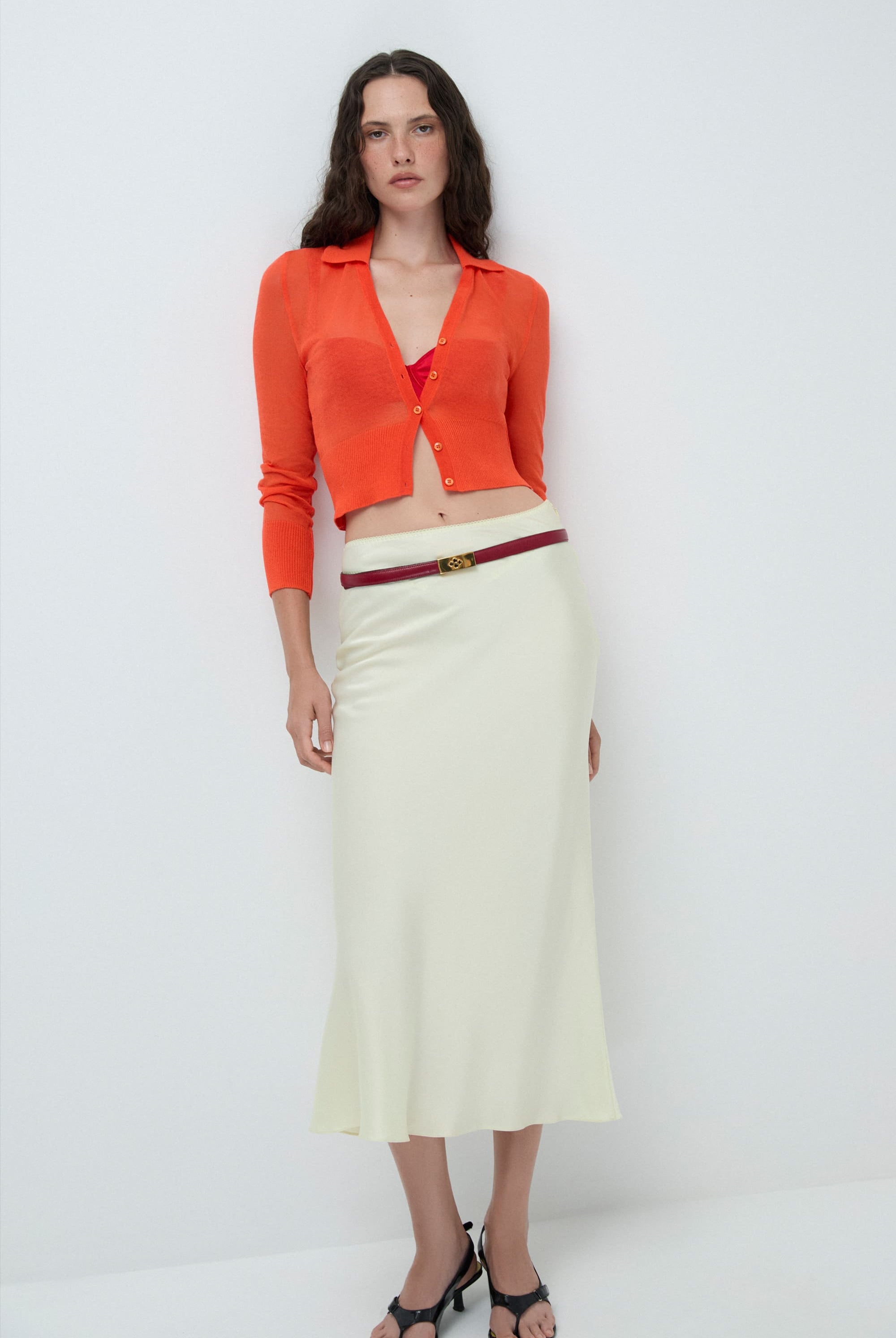 Woman wearing an orange Cecilia cardigan white skirt with belt on a white background, front view