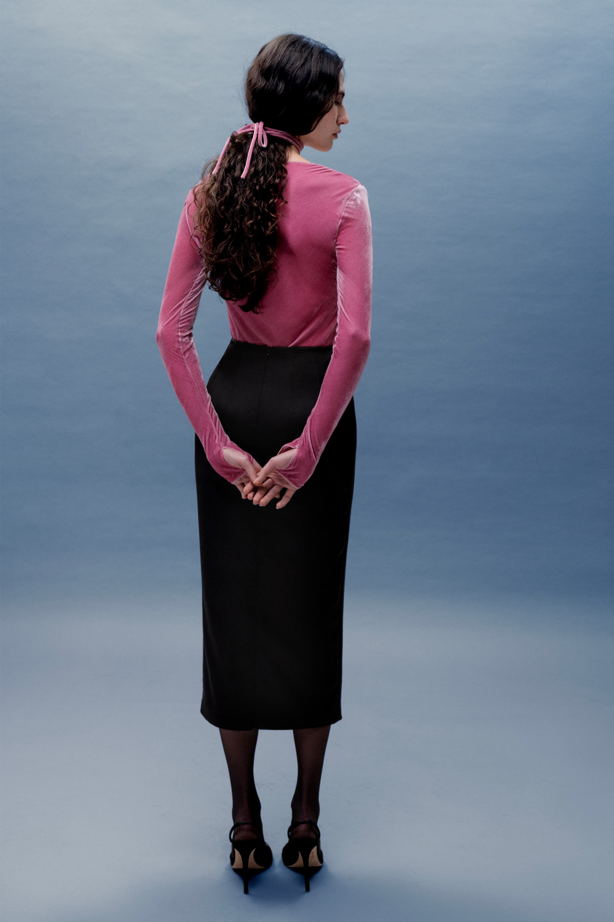 Woman wearing a pink long-sleeve Massima top and black skirt against a gray background