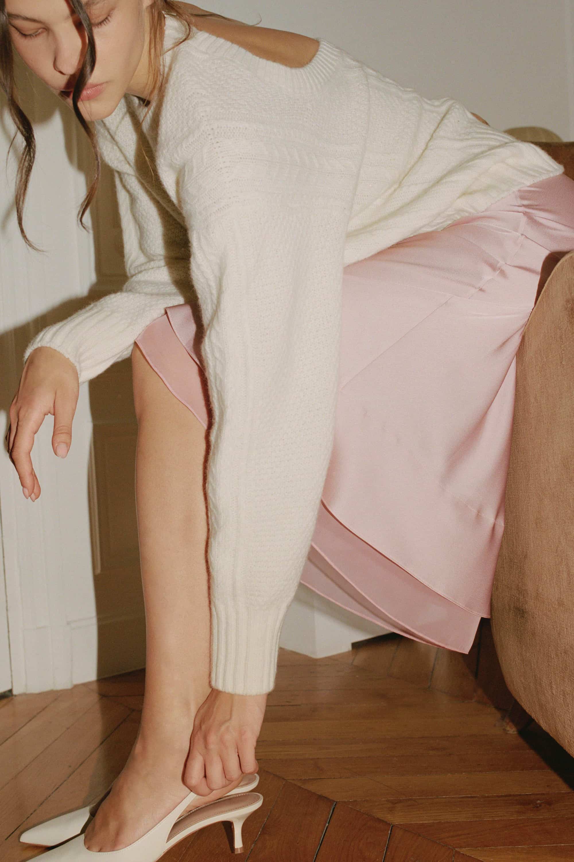 Woman in a ivory Josie cardigan and powder pink Elina skirt bent down to adjust her shoe