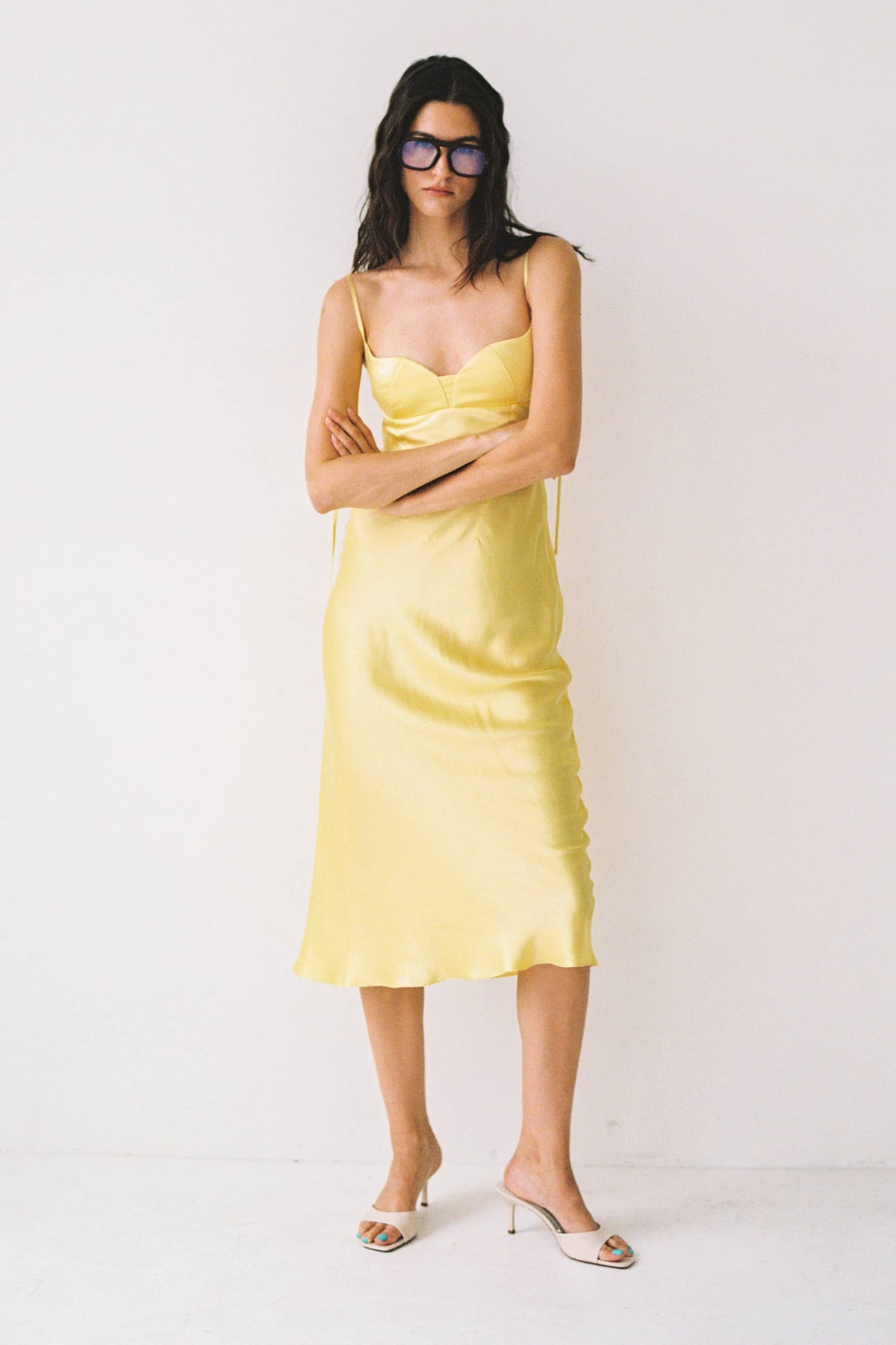 A woman modeling a pale yellow satin slip Isolde dress with thin straps, paired with white heeled mule sandals and dark sunglasses, against a plain white studio background