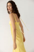 A woman modeling a pale yellow satin slip Isolde dress with thin spaghetti straps and a tie-back detail, photographed against a clean white studio background, side view