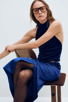 A woman wearing a sleeveless dark blue velvet mock-neck top and matching blue skirt sits on a wooden stool against a light gray background.