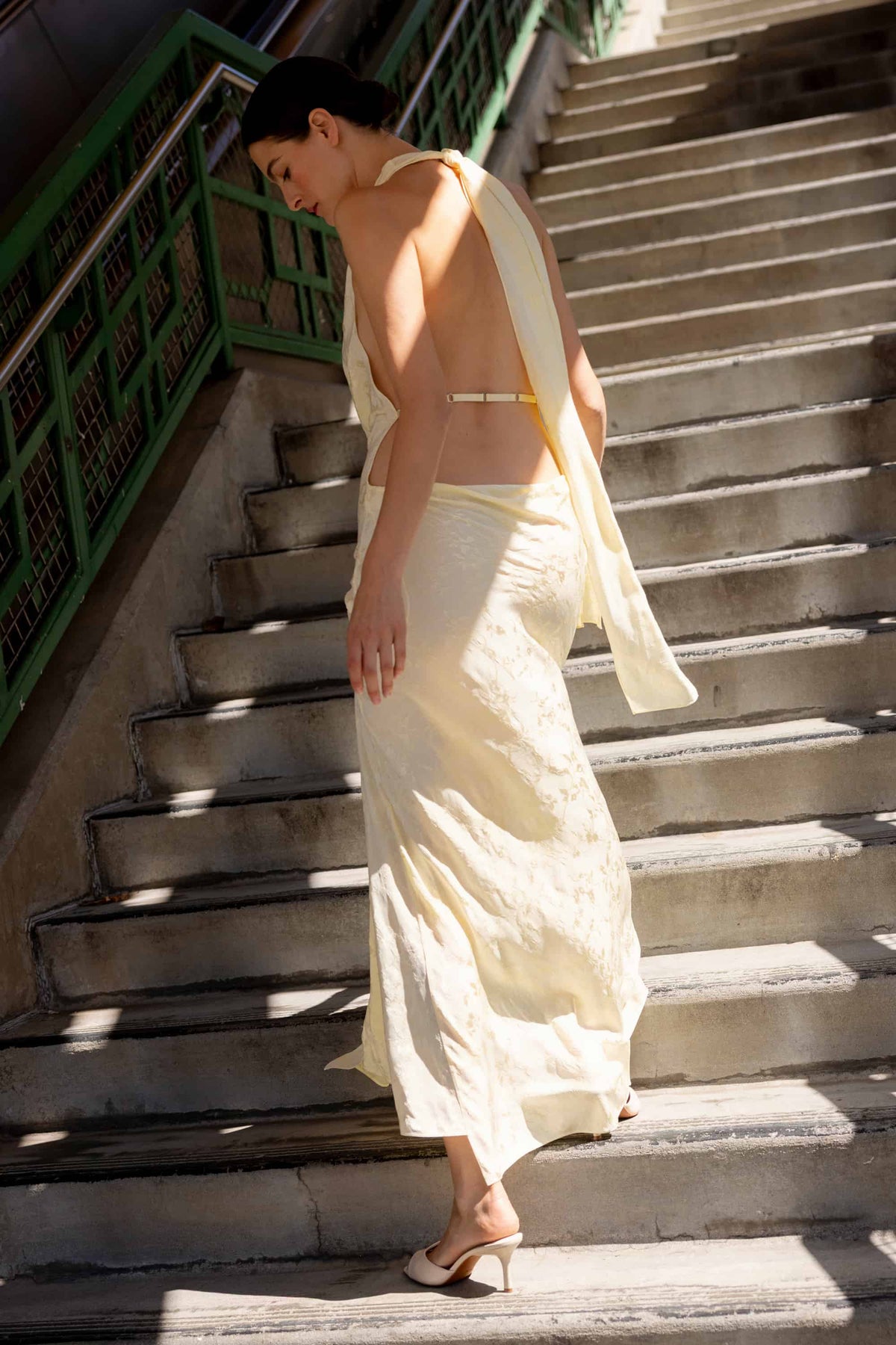 A woman wearing a draped buttermilk-colored maxi Kelly dress with an open back and halter-style shoulder detail, walking down outdoor concrete stairs with green railings