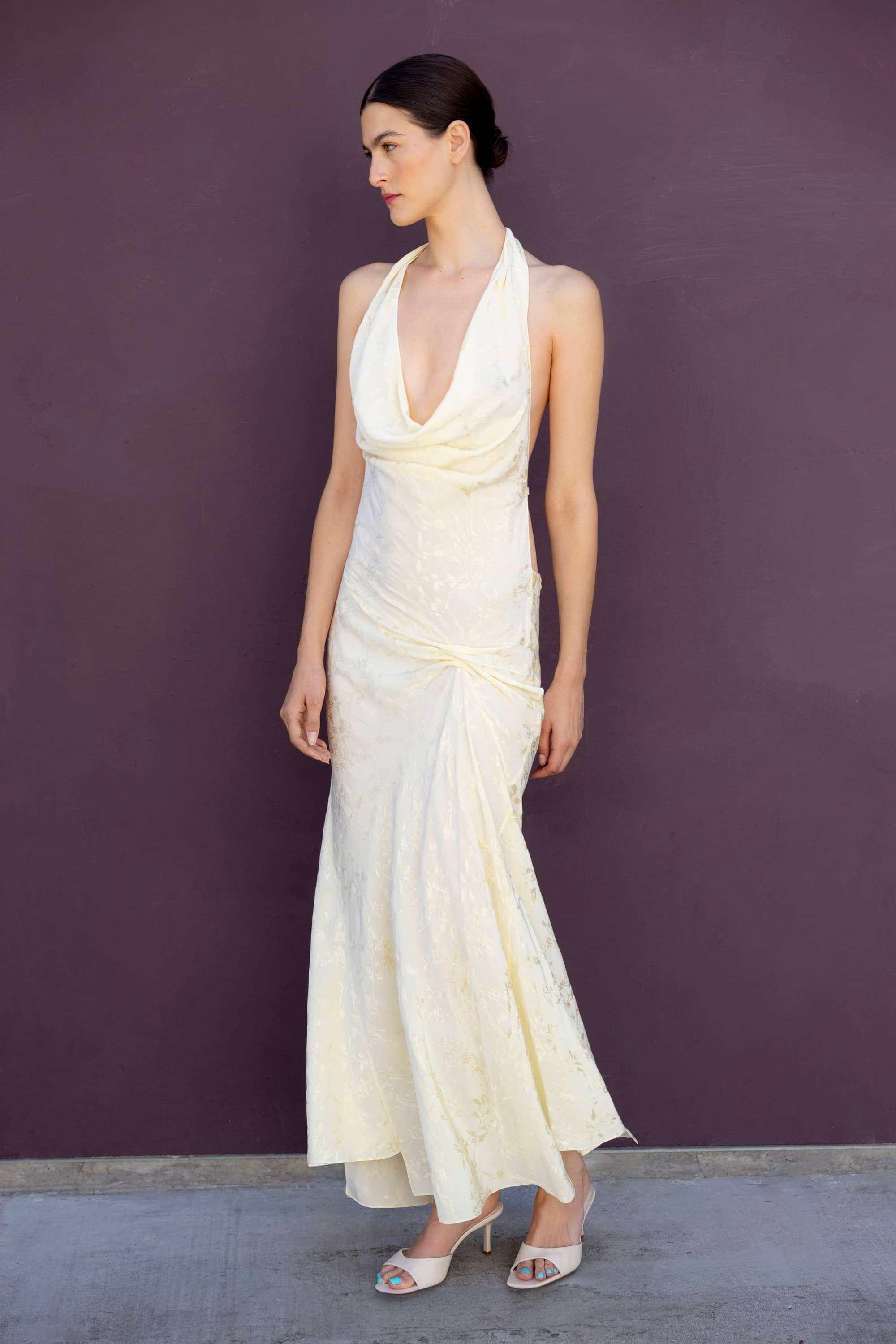 A woman modeling a buttermilk-colored halter-neck maxi Kelly dress with a draped, gathered design and a flared skirt, standing against a solid dark purple wall