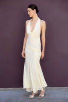 A woman modeling a buttermilk-colored halter-neck maxi Kelly dress with a draped, gathered design and a flared skirt, standing against a solid dark purple wall