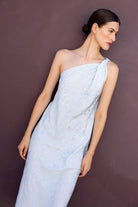 A woman wearing a light blue one-shoulder Rina dress with a textured, shimmering fabric, posed against a dark, textured brown wall