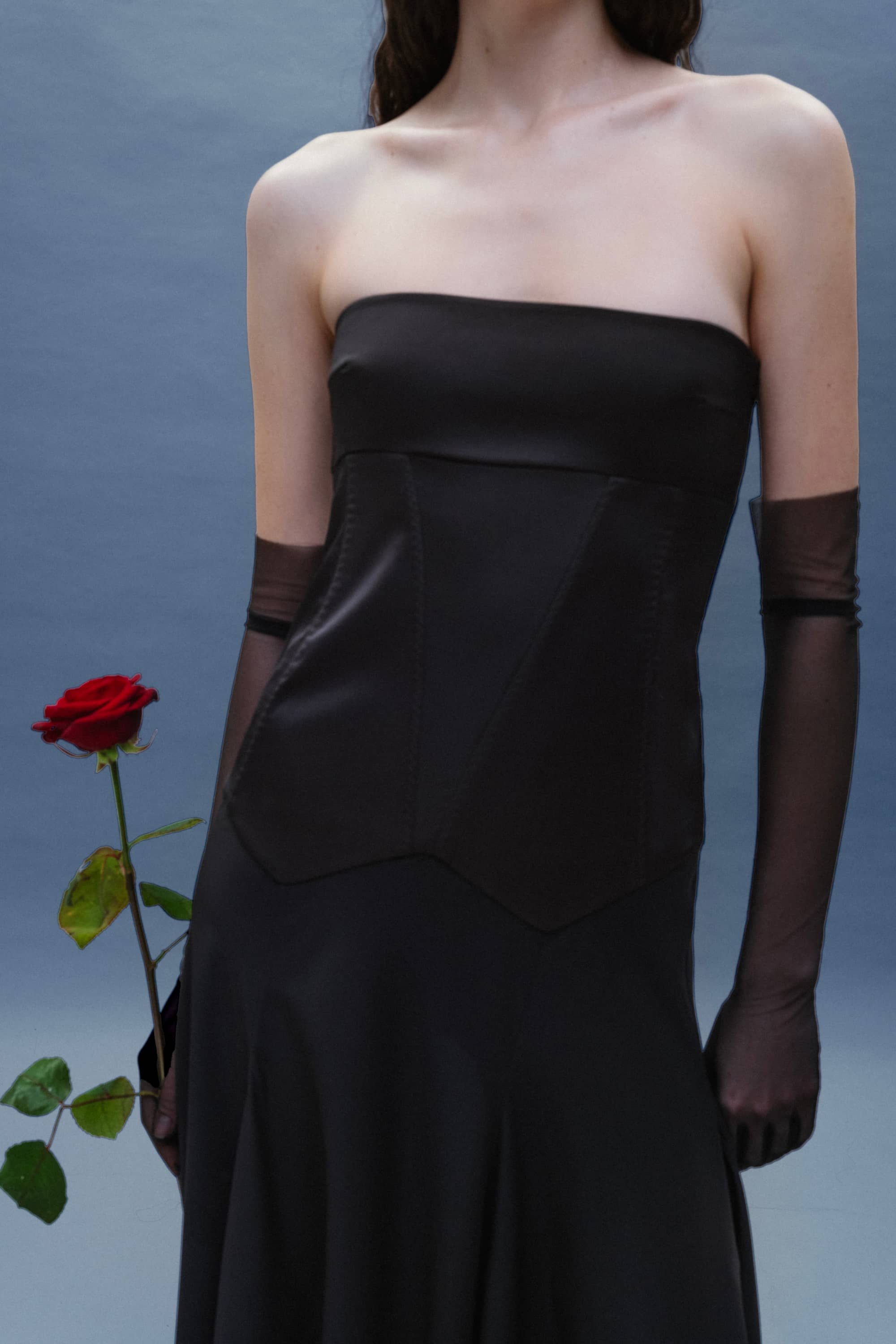 Woman in a black strapless Ariella dress holding a red rose against a gray background