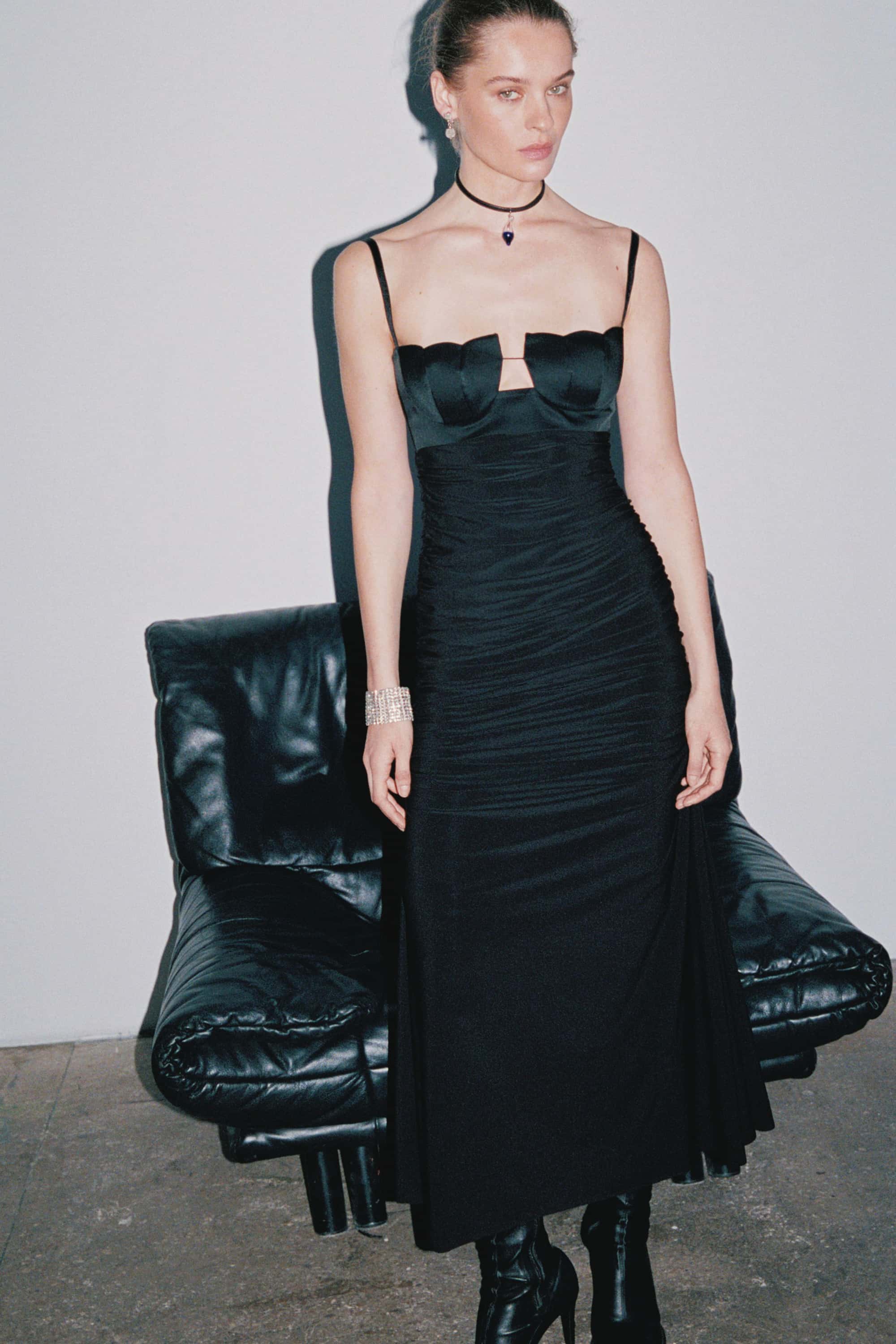 Woman wearing a black Vivian dress standing next to a black leather chair against a plain background