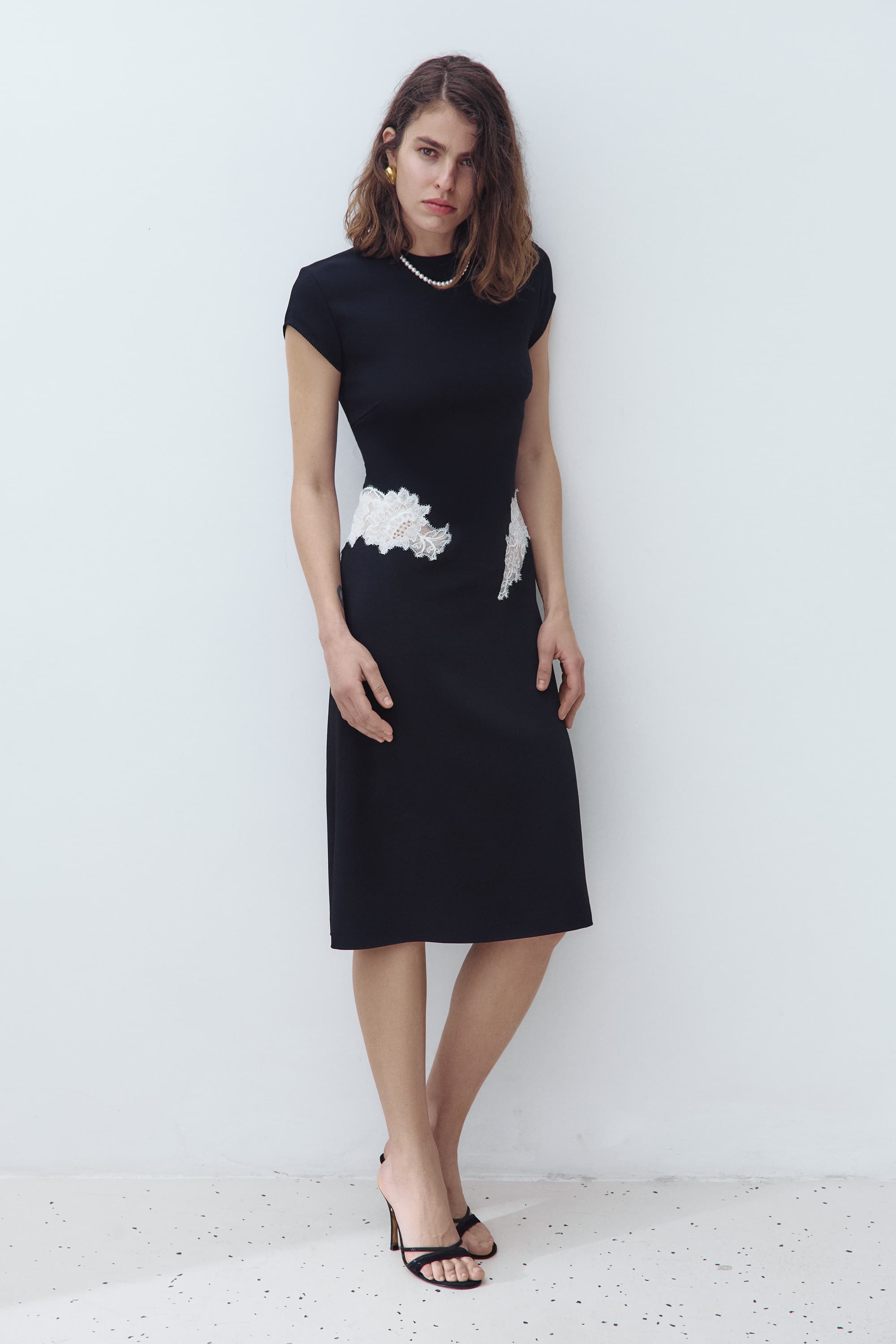 A woman modeling a black knee-length Eleni dress with short cap sleeves and a white textured floral applique detail at the waist, standing against a plain light gray wall