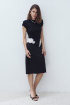 A woman modeling a black knee-length Eleni dress with short cap sleeves and a white textured floral applique detail at the waist, standing against a plain light gray wall