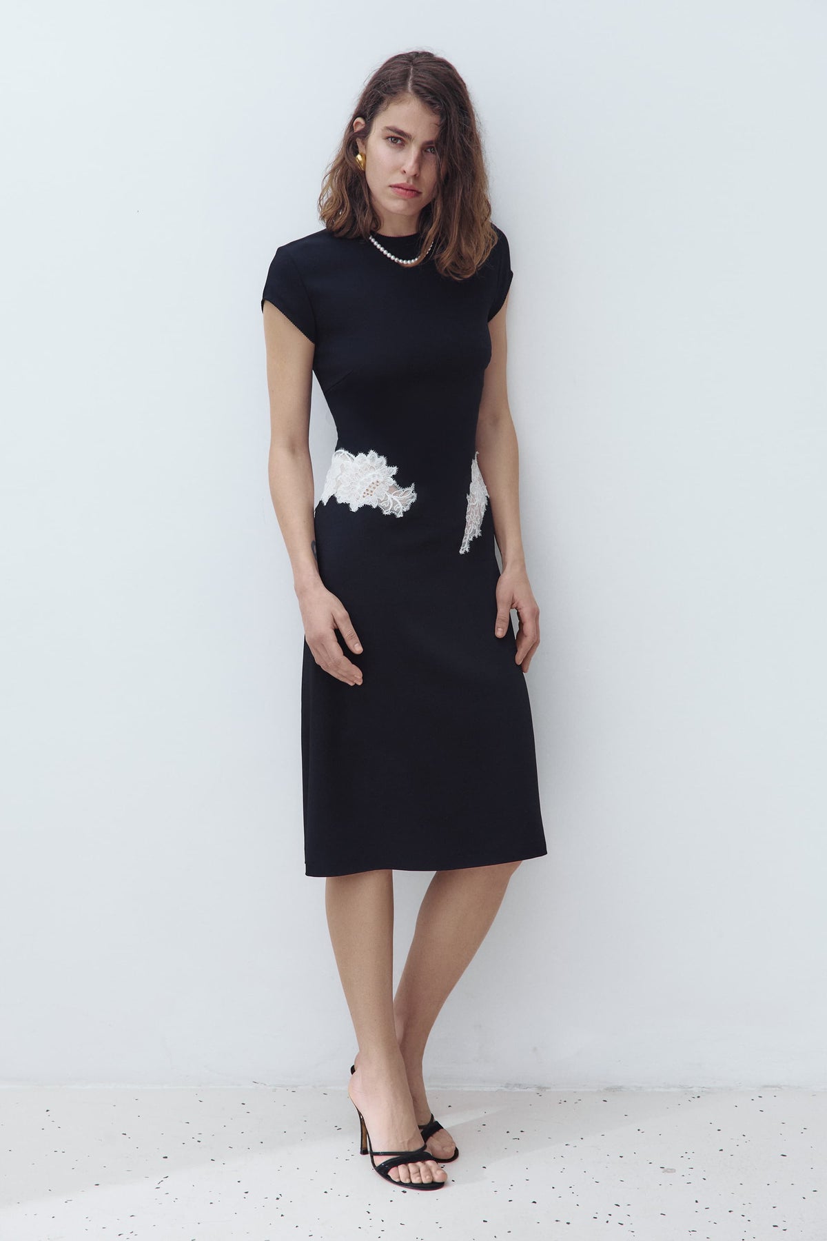 A woman modeling a black knee-length Eleni dress with short cap sleeves and a white textured floral applique detail at the waist, standing against a plain light gray wall
