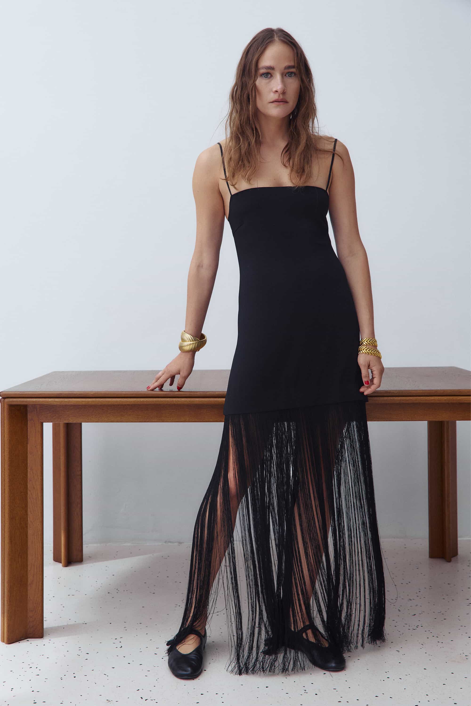 A woman models a black spaghetti-strap maxi Evangelia dress with a solid upper bodice and a sheer, fringed tulle hem, standing next to a wooden table against a white wall