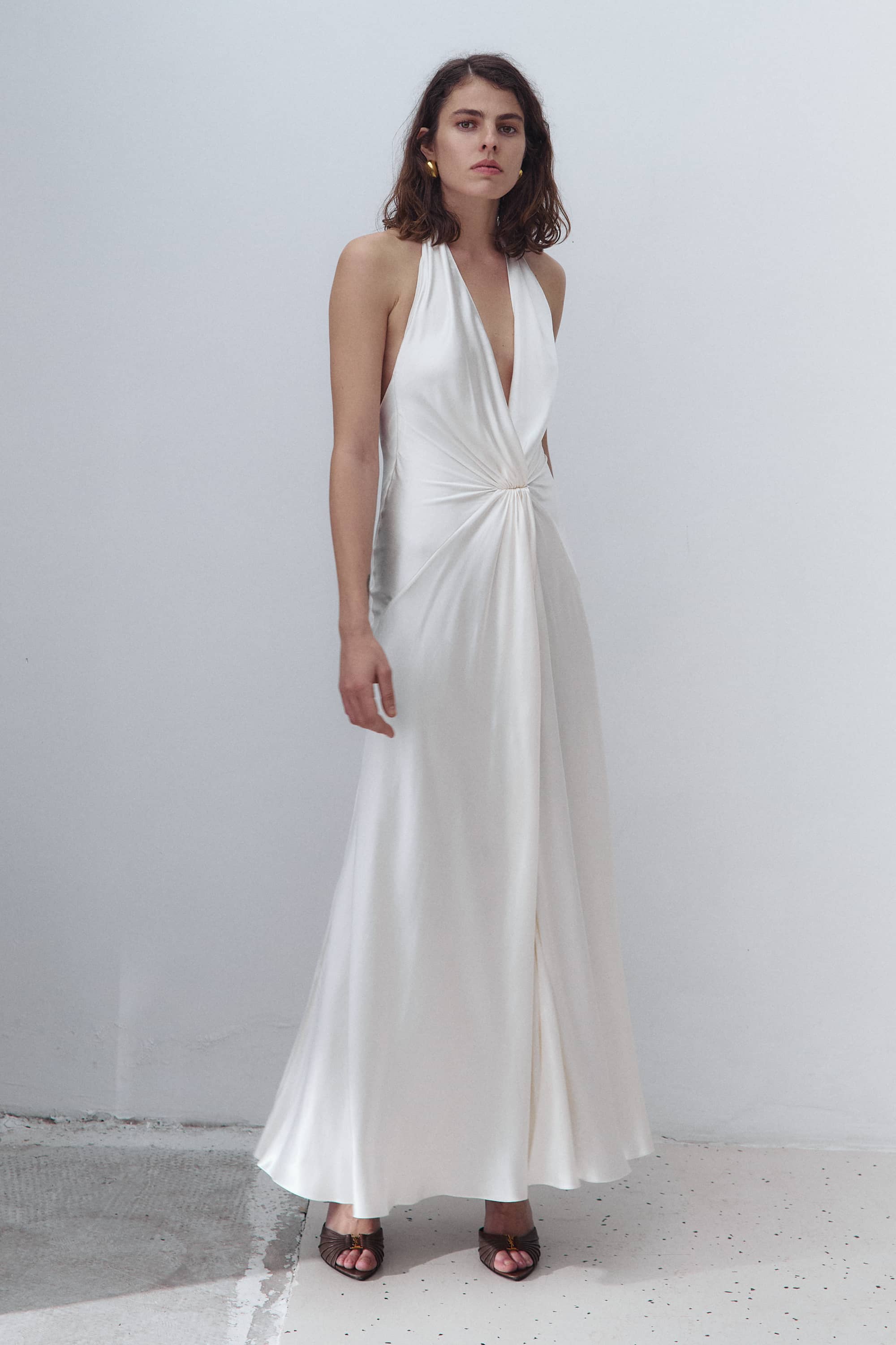 A woman modeling a floor-length white halter Ruth dress with a deep V-neckline and a gathered twist detail at the waist, standing against a plain light gray wall