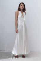A woman modeling a floor-length white halter Ruth dress with a deep V-neckline and a gathered twist detail at the waist, standing against a plain light gray wall