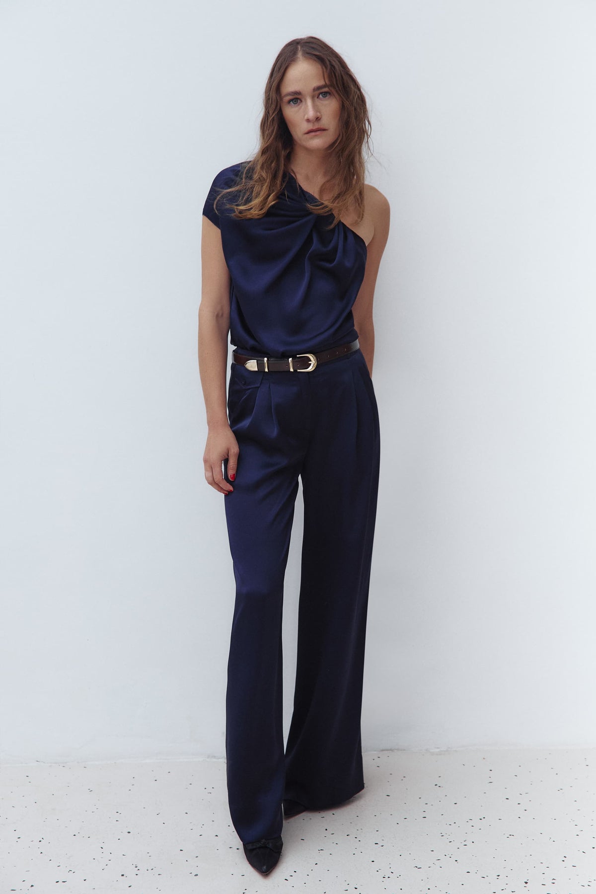 A woman modeling a dark navy blue one-shoulder draped Paris top and matching navy wide-leg Katerina trousers, styled with a brown belt and gold bracelets against a light gray studio background