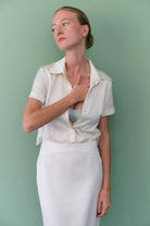 Woman wearing a white Arisa dress with a slit skirt and black belt against a light green background, front view