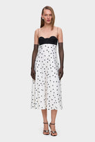 Woman wearing a black and white polka dot Erika dress with thin straps and black gloves on a white background, front view