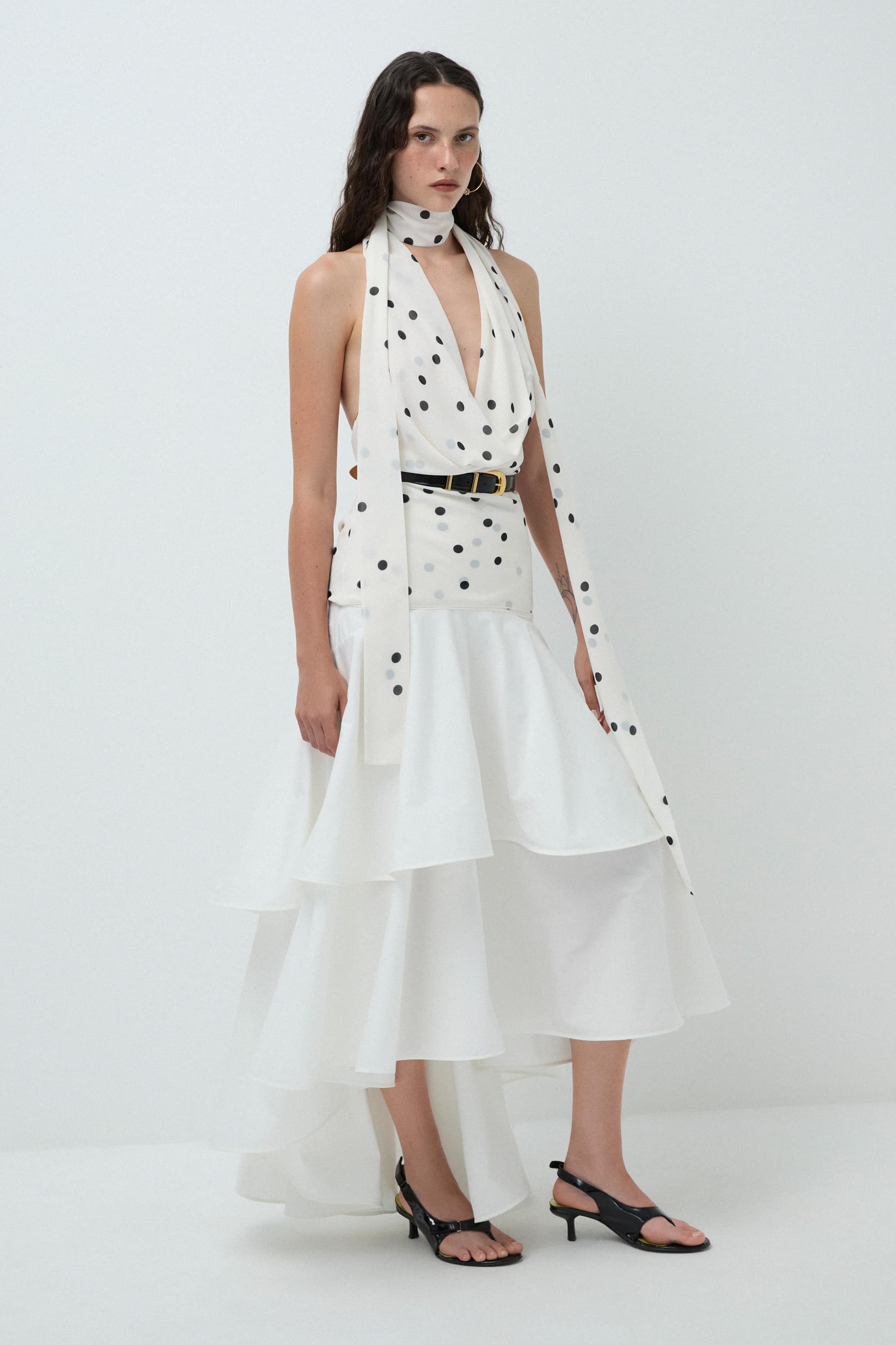 Woman wearing a white sleeveless Agnes top with black polka dots and a belt on a white background, side view